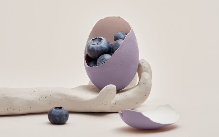 An outstretched hand made of clay, holding a purple egg whose shell has been cracked to reveal "superfood" blueberries inside.