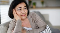 a sixty-something woman sits on the couch at home looking thoughtful as she wonders if a younger therapist could really help her