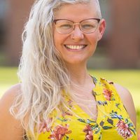 Dr. Emily K. Sandoz smiling in a bright yellow floral shirt with her silvery hair swept over one shoulder.