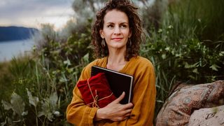 Woman journalling and holding fabric wrapped gift as she sits on the shores of a lake