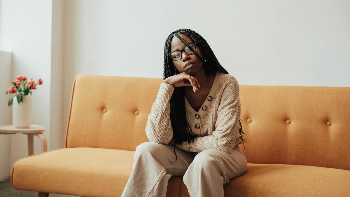 A Black female therapist with box braids sits on a midcentury sofa upholstered in tangerine linen.