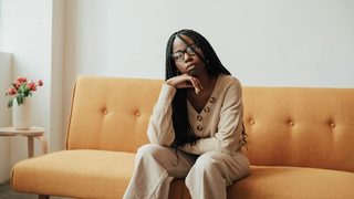 A Black female therapist with box braids sits on a midcentury sofa upholstered in tangerine linen.
