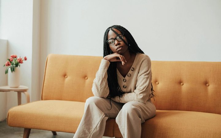 A Black female therapist with box braids sits on a midcentury sofa upholstered in tangerine linen.