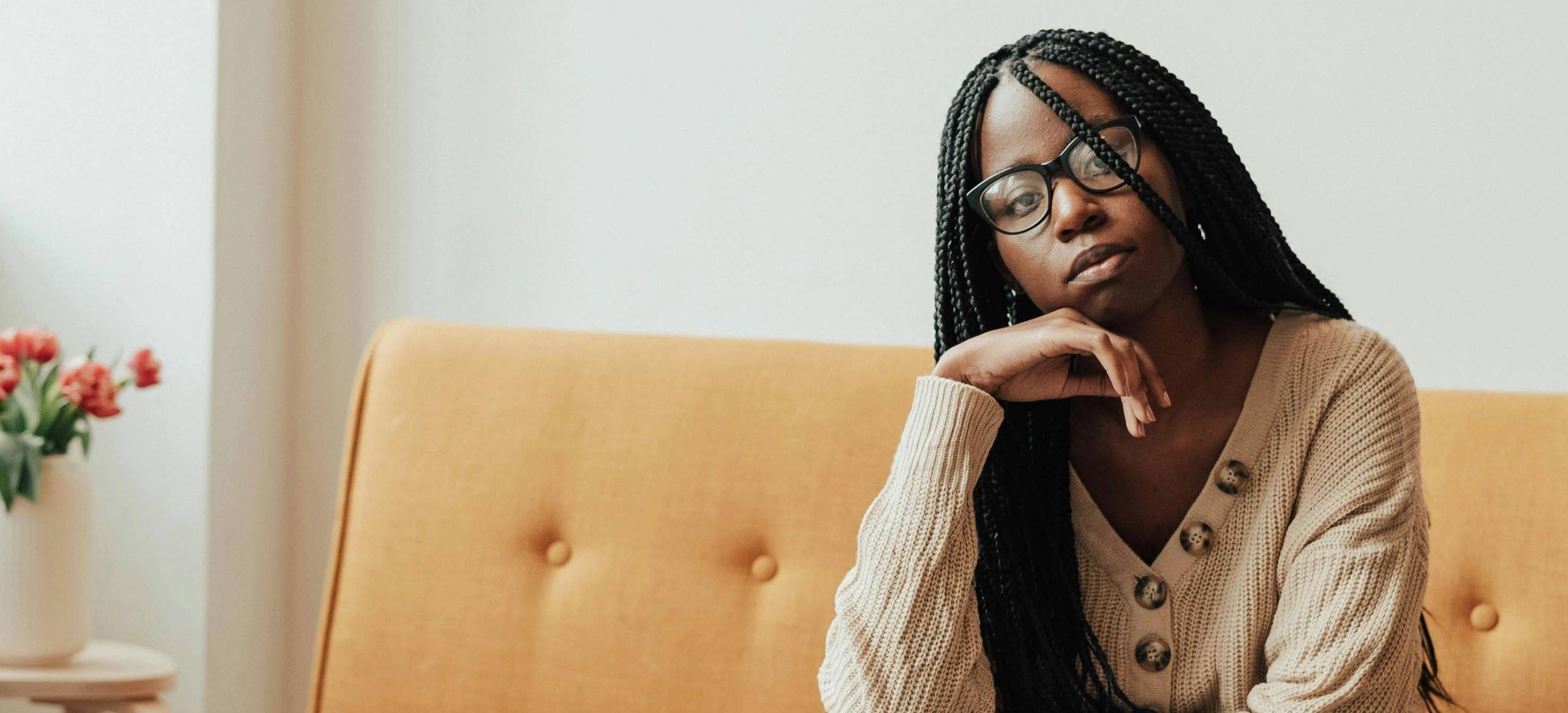 A Black woman with long braids sitting on a sofa, resting her chin on her hand