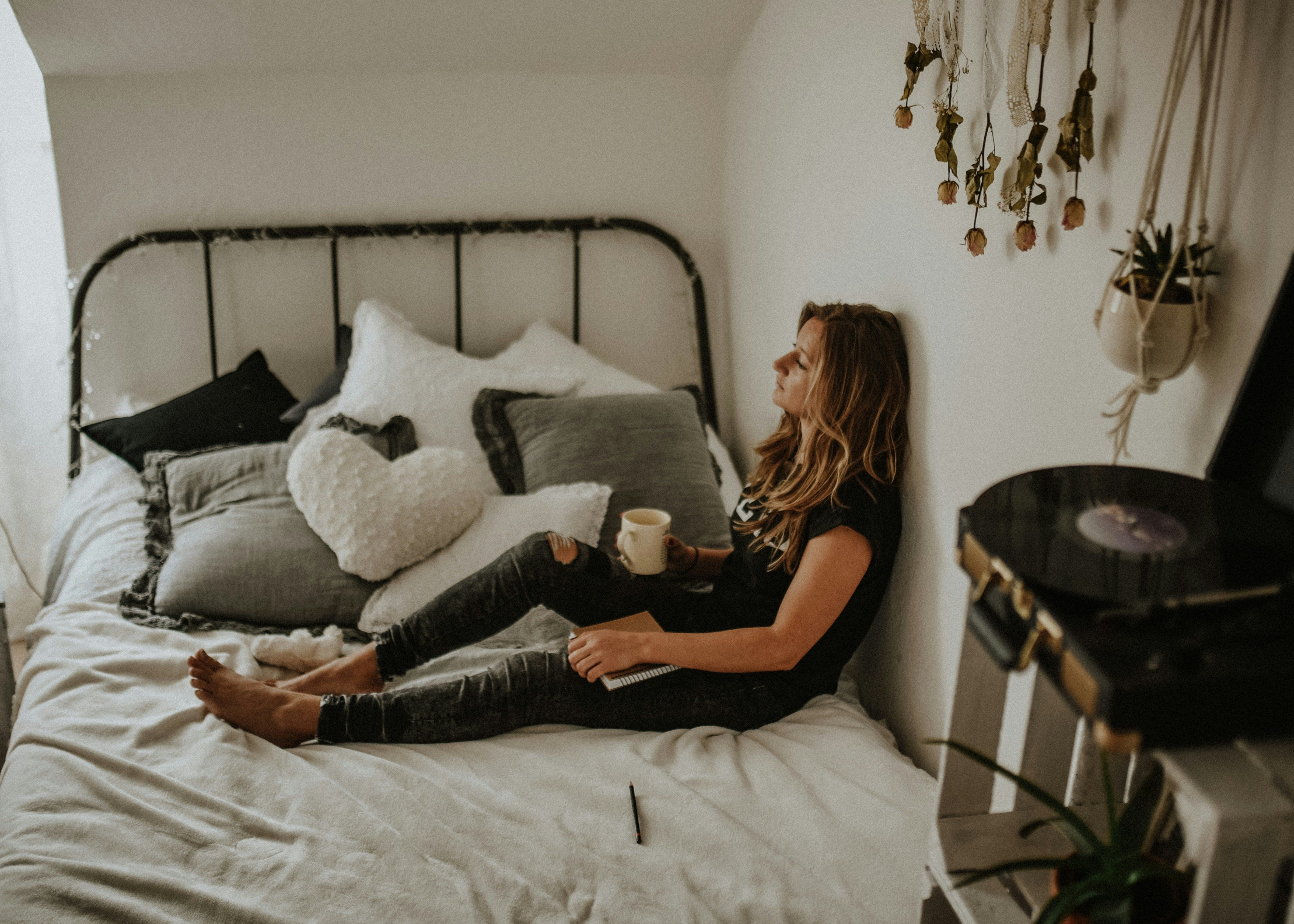 A person lying in bed with their back against their wall, holding a coffee mug in one hand and a journal in the other, with their legs outstretched. They are surrounded by hanging plants, a nearby record player, and a mountain of pillows. They are staring in the direction of a window with a thoughtful expression.