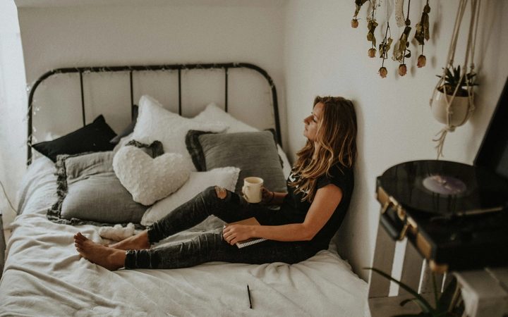 A person lying in bed with their back against their wall, holding a coffee mug in one hand and a journal in the other, with their legs outstretched. They are surrounded by hanging plants, a nearby record player, and a mountain of pillows. They are staring in the direction of a window with a thoughtful expression.