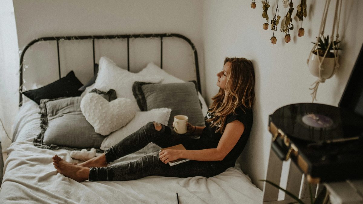 A person lying in bed with their back against their wall, holding a coffee mug in one hand and a journal in the other, with their legs outstretched. They are surrounded by hanging plants, a nearby record player, and a mountain of pillows. They are staring in the direction of a window with a thoughtful expression.