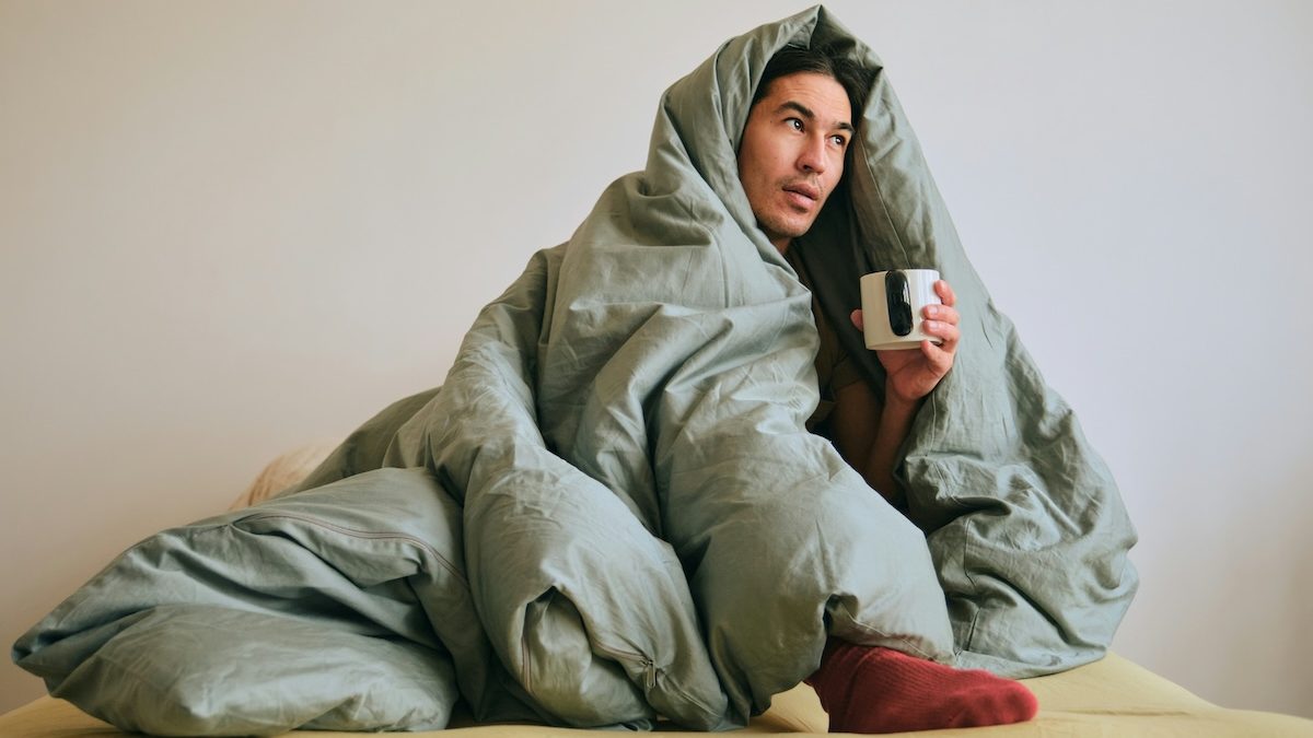 A man dealing with seasonal affective disorder is huddling in bed, wrapped in a duvet and drinking coffee