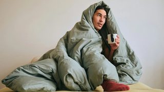 A man dealing with seasonal affective disorder is huddling in bed, wrapped in a duvet and drinking coffee