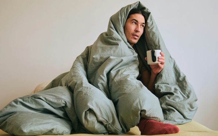 A man dealing with seasonal affective disorder is huddling in bed, wrapped in a duvet and drinking coffee