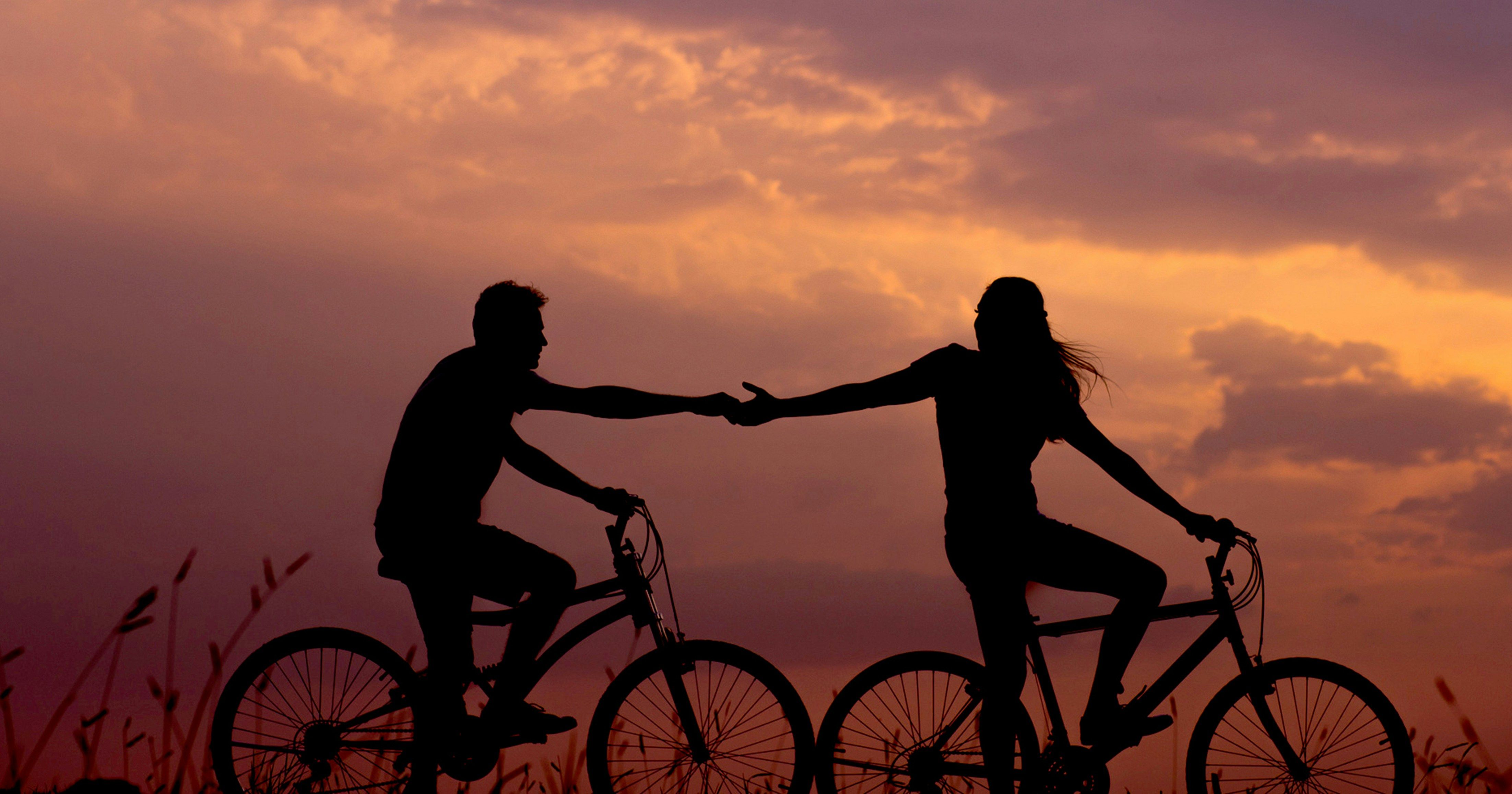 Silhouettes of two people on bicycles reaching for each other at sunset.