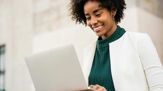 A mental health therapist in a green top and white suit jacket is sitting outside with her laptop, smiling as she updates her directory profile