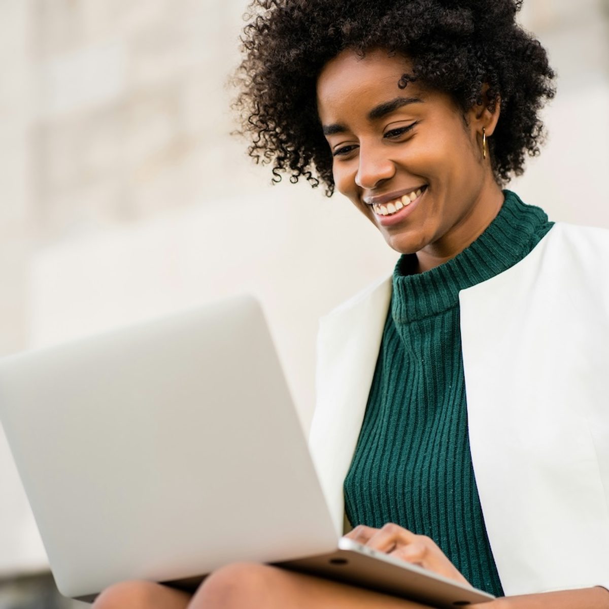 A mental health therapist in a green top and white suit jacket is sitting outside with her laptop, smiling as she updates her directory profile