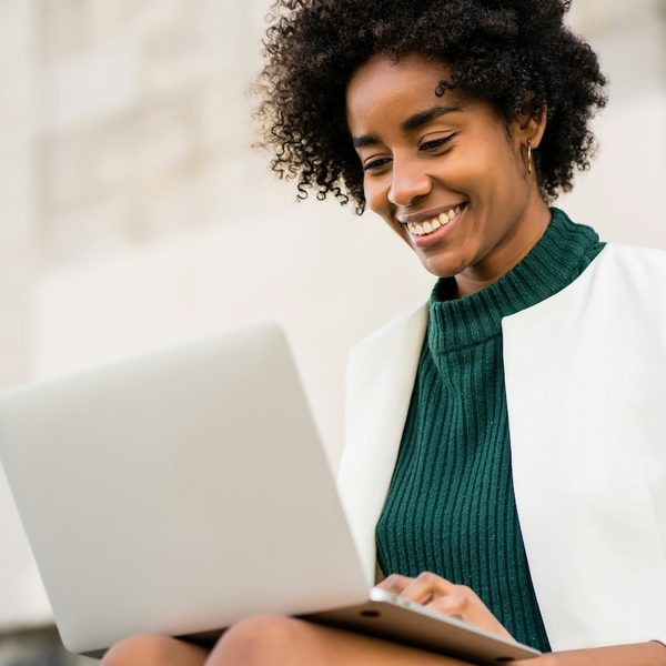 A mental health therapist in a green top and white suit jacket is sitting outside with her laptop, smiling as she updates her directory profile