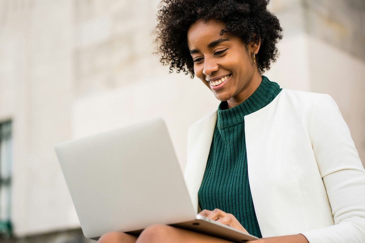A mental health therapist in a green top and white suit jacket is sitting outside with her laptop, smiling as she updates her directory profile