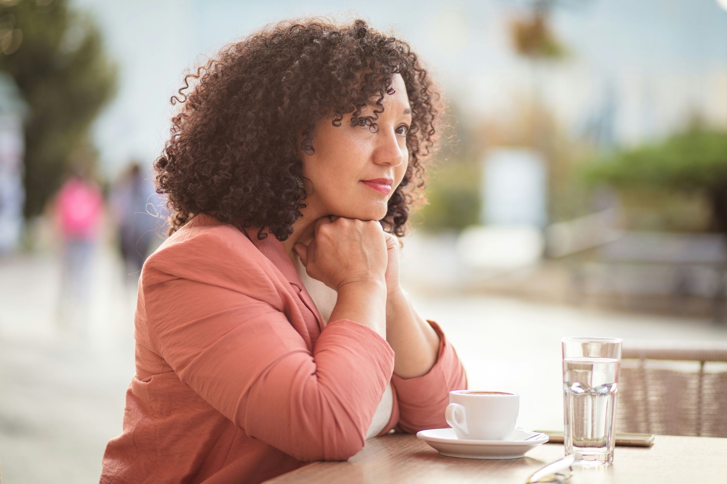 A woman with black, curly hair wearing a salmon colored blazer sitting alone at a cafe. Her chin is resting on her hands and she looks thoughtful and genuinely concerned.