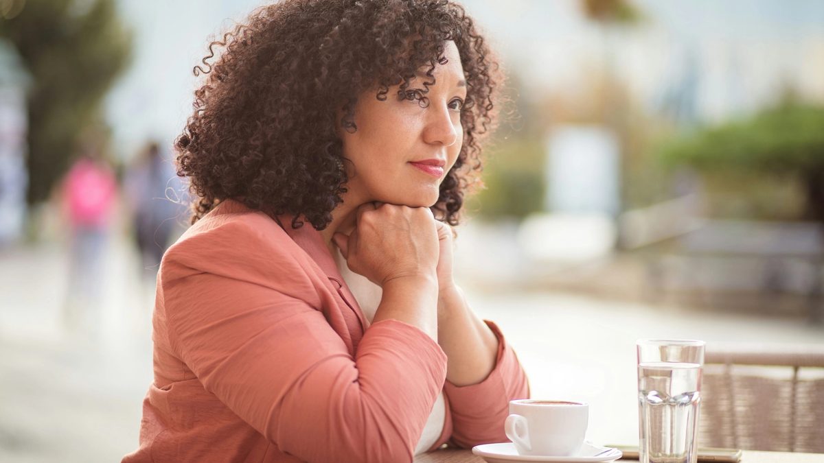 A woman with black, curly hair wearing a salmon colored blazer sitting alone at a cafe. Her chin is resting on her hands and she looks thoughtful and genuinely concerned.