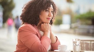 A woman with black, curly hair wearing a salmon colored blazer sitting alone at a cafe. Her chin is resting on her hands and she looks thoughtful and genuinely concerned.