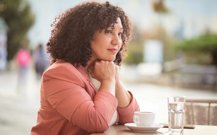 A woman with black, curly hair wearing a salmon colored blazer sitting alone at a cafe. Her chin is resting on her hands and she looks thoughtful and genuinely concerned.