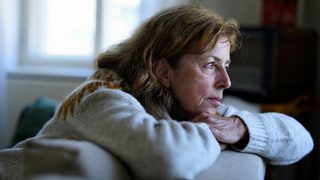 An older woman sits leaning on the back of a couch, missing her adult child