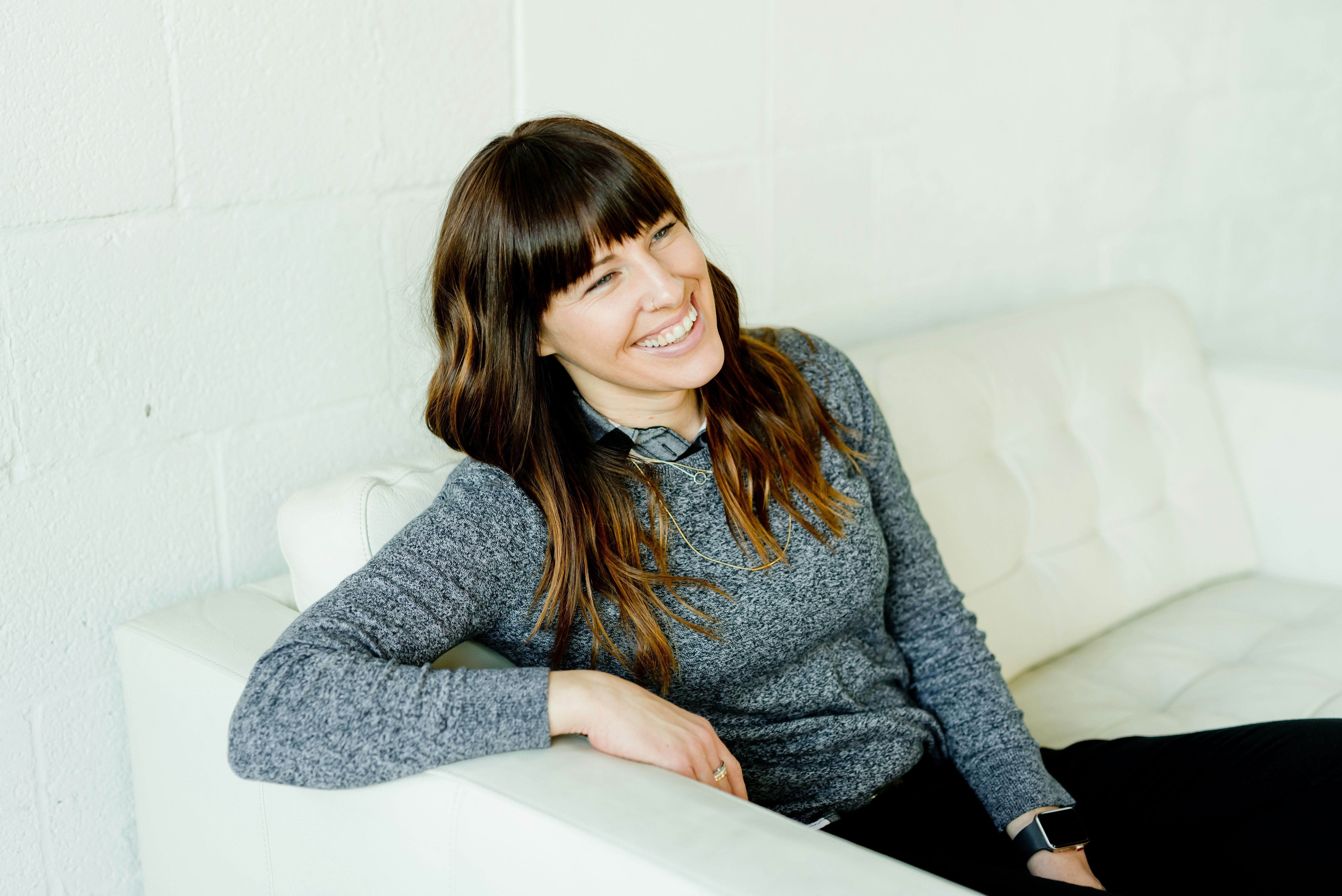 A smiling person with long brown hair sits reclined on a white sofa, leaning on one arm, gazing across the room.