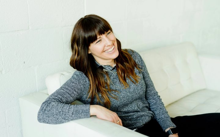 A smiling person with long brown hair sits reclined on a white sofa, leaning on one arm, gazing across the room.