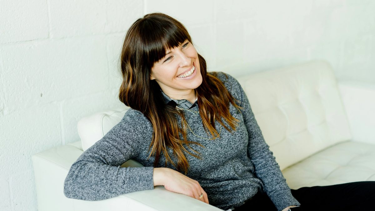 A smiling person with long brown hair sits reclined on a white sofa, leaning on one arm, gazing across the room.