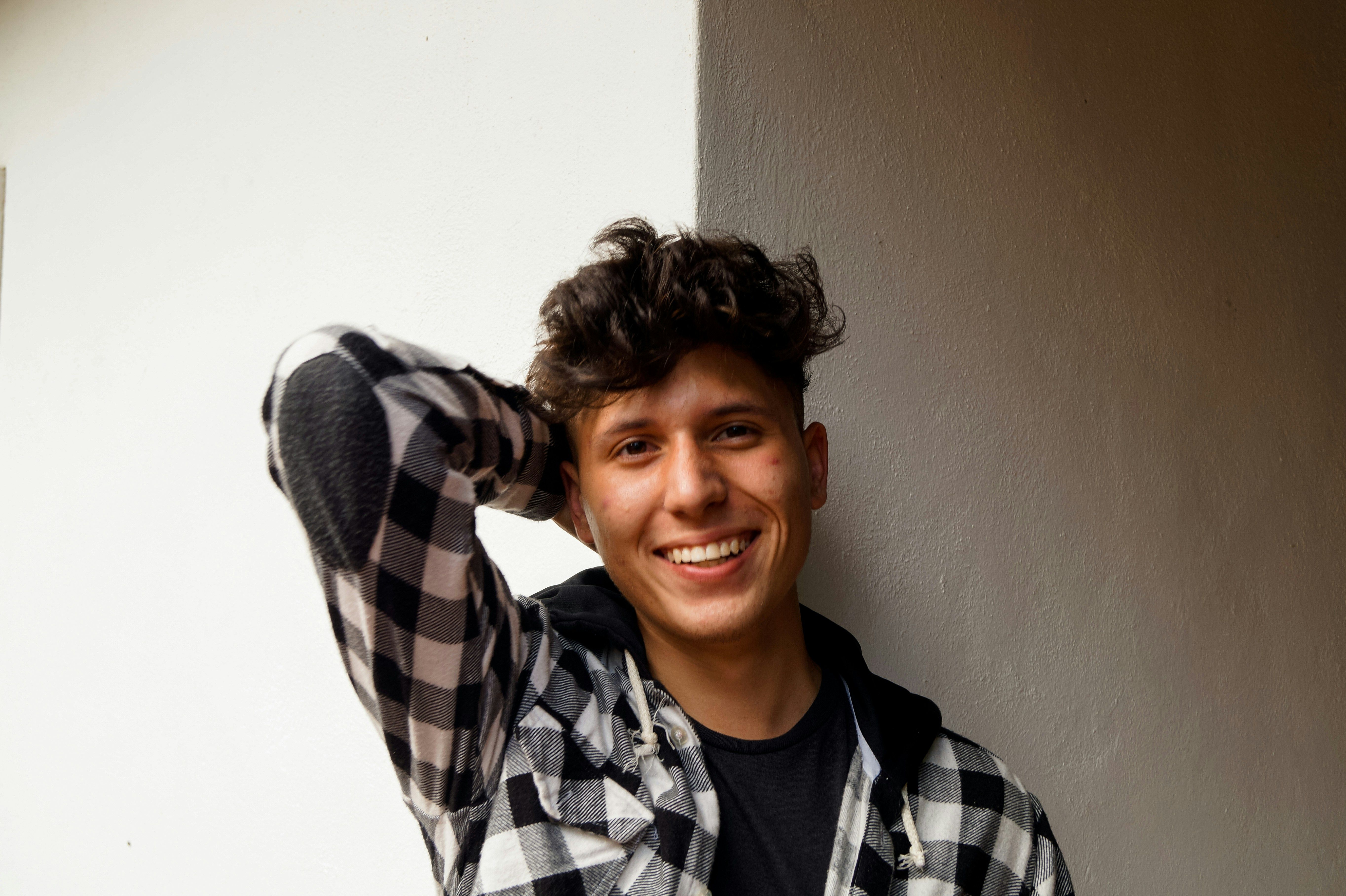 A smiling, masculine individual with light skin gazes into the camera with one arm folded behind their head. They wear a black and white checkered shirt over a dark tee. Photo by Jorge Salvador via Unsplash.