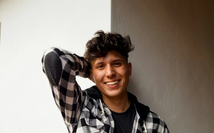 A smiling, masculine individual with light skin gazes into the camera with one arm folded behind their head. They wear a black and white checkered shirt over a dark tee. Photo by Jorge Salvador via Unsplash.