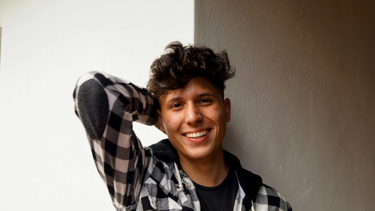 A smiling, masculine individual with light skin gazes into the camera with one arm folded behind their head. They wear a black and white checkered shirt over a dark tee. Photo by Jorge Salvador via Unsplash.