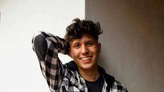 A smiling, masculine individual with light skin gazes into the camera with one arm folded behind their head. They wear a black and white checkered shirt over a dark tee. Photo by Jorge Salvador via Unsplash.
