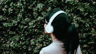 A person with a dark ponytail and white headphones stands facing a leafy bush.