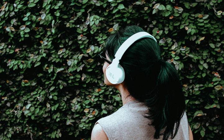 A person with a dark ponytail and white headphones stands facing a leafy bush.
