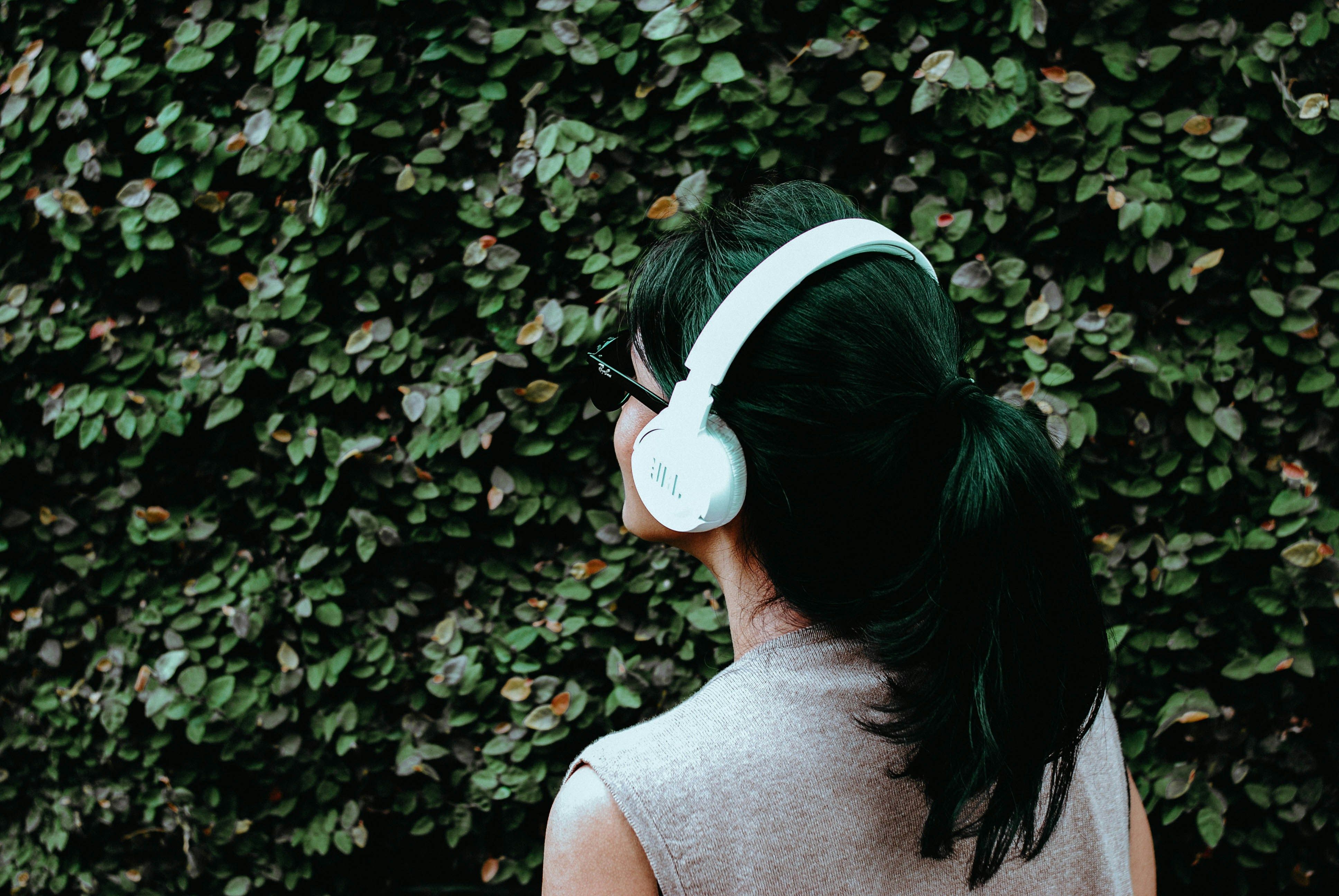 A person with a dark ponytail and white headphones stands facing a leafy bush.