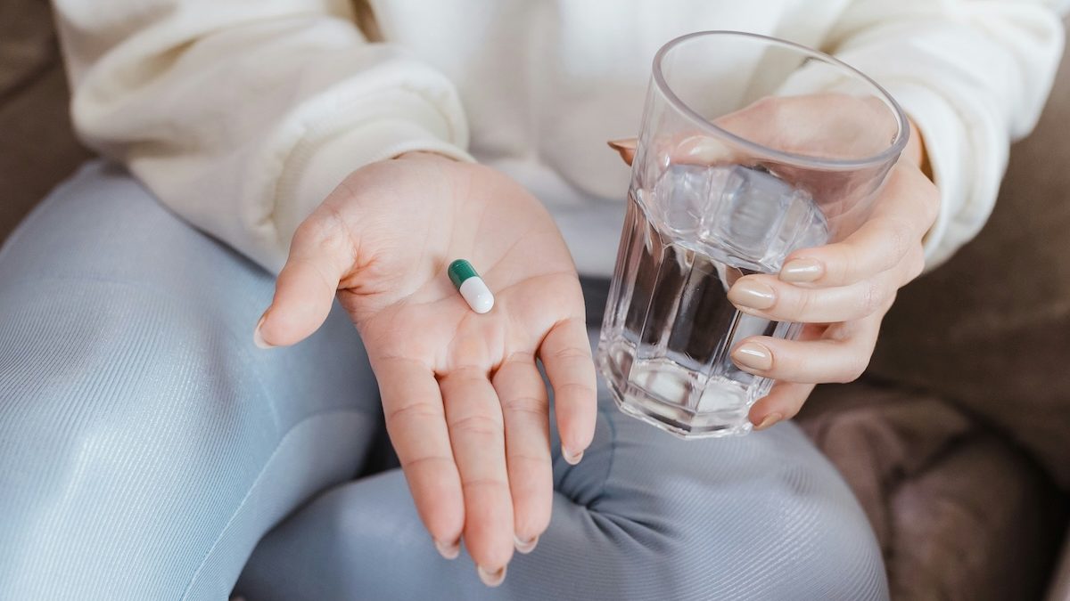 A close-up of a seated woman holding a pill in one hand and a glass of water in the other