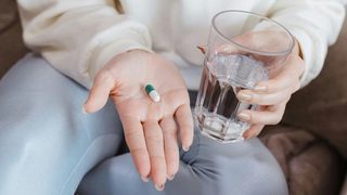 A close-up of a seated woman holding a pill in one hand and a glass of water in the other