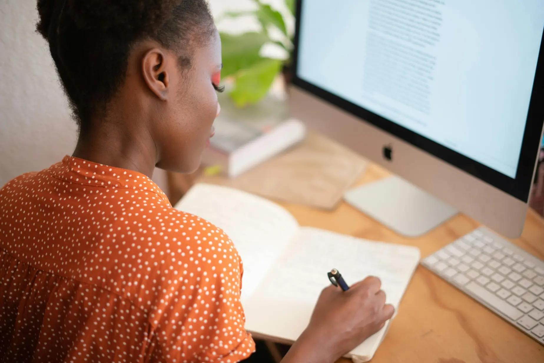 Person sitting in front of a computer and writing notes in a notebook on the desk