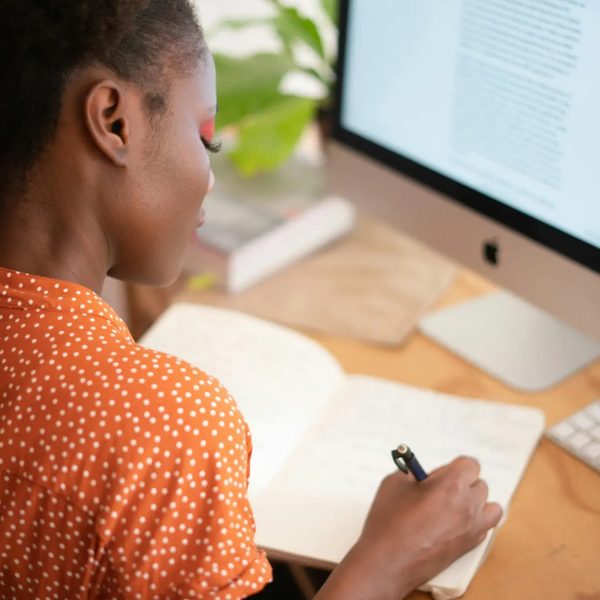 Person sitting in front of a computer and writing notes in a notebook on the desk
