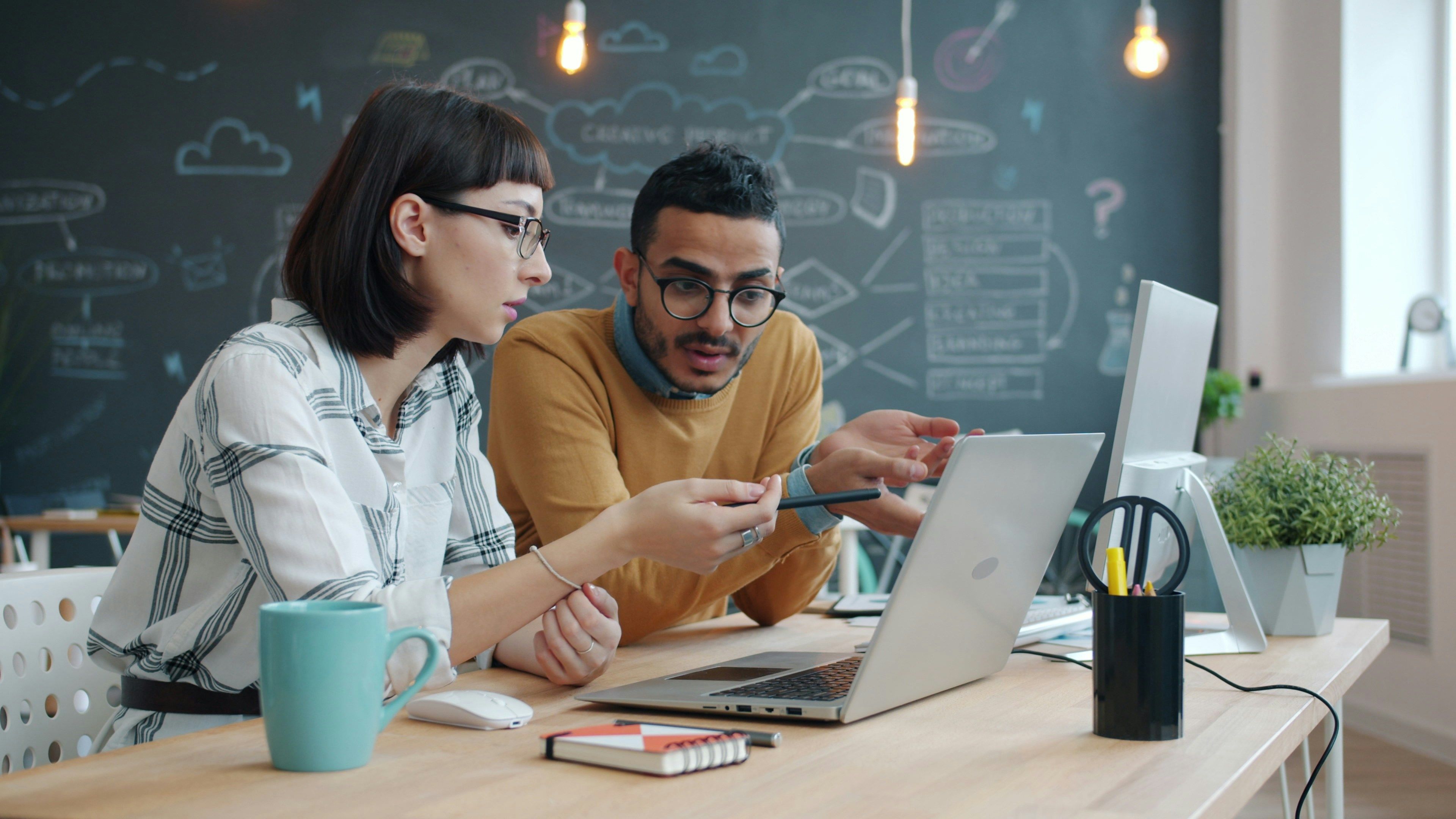 Two coworkers discussing work at a laptop in a creative office.