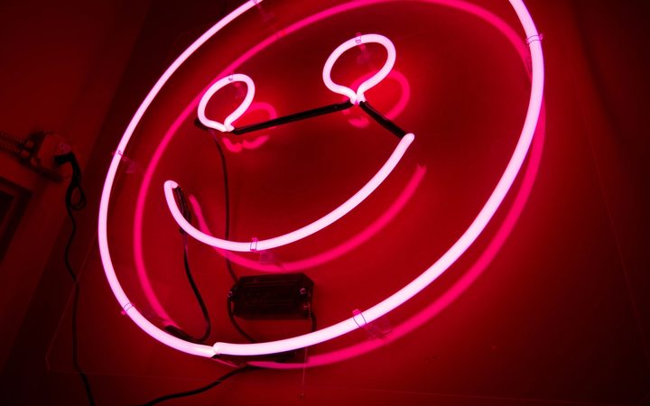 A neon pink sign in the shape of a smiley face looks down on a dark room, imposing its toxic positivity on all.