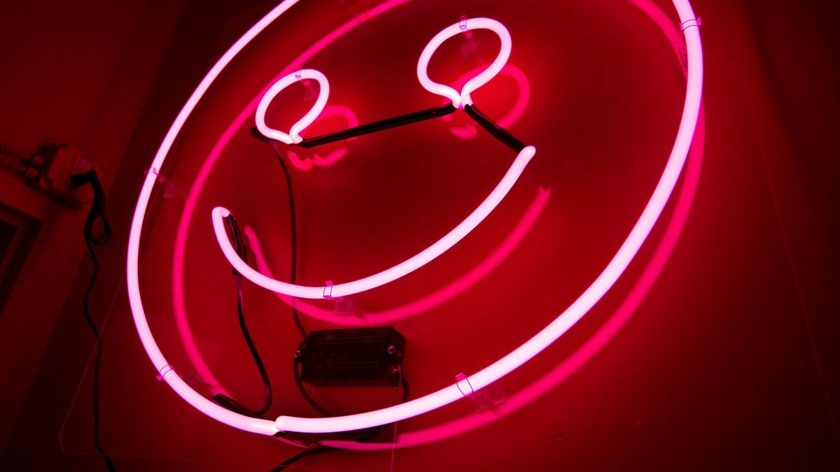 A neon pink sign in the shape of a smiley face looks down on a dark room, imposing its toxic positivity on all.