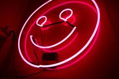 A neon pink sign in the shape of a smiley face looks down on a dark room, imposing its toxic positivity on all.