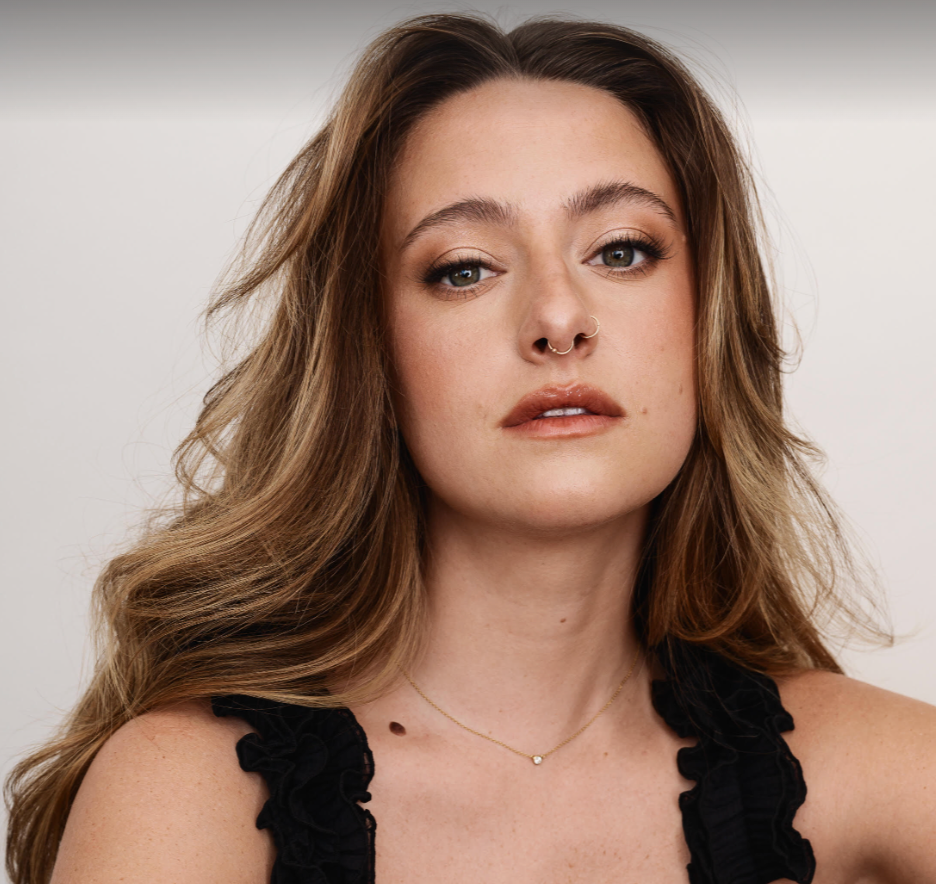 Haley Jakobson, a white femme with long brown wavy hair gazes into the camera with a curious gaze.