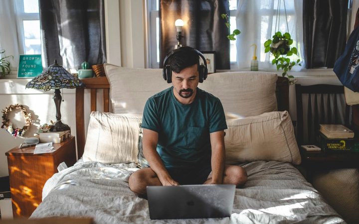 A masculine-appearing person sits cross-legged in bed looking down at a laptop, with headphones on. They are in a cluttered but cozy room, with plants, bright windows, and decorative items.