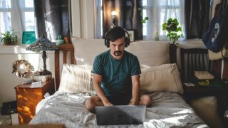 A masculine-appearing person sits cross-legged in bed looking down at a laptop, with headphones on. They are in a cluttered but cozy room, with plants, bright windows, and decorative items.