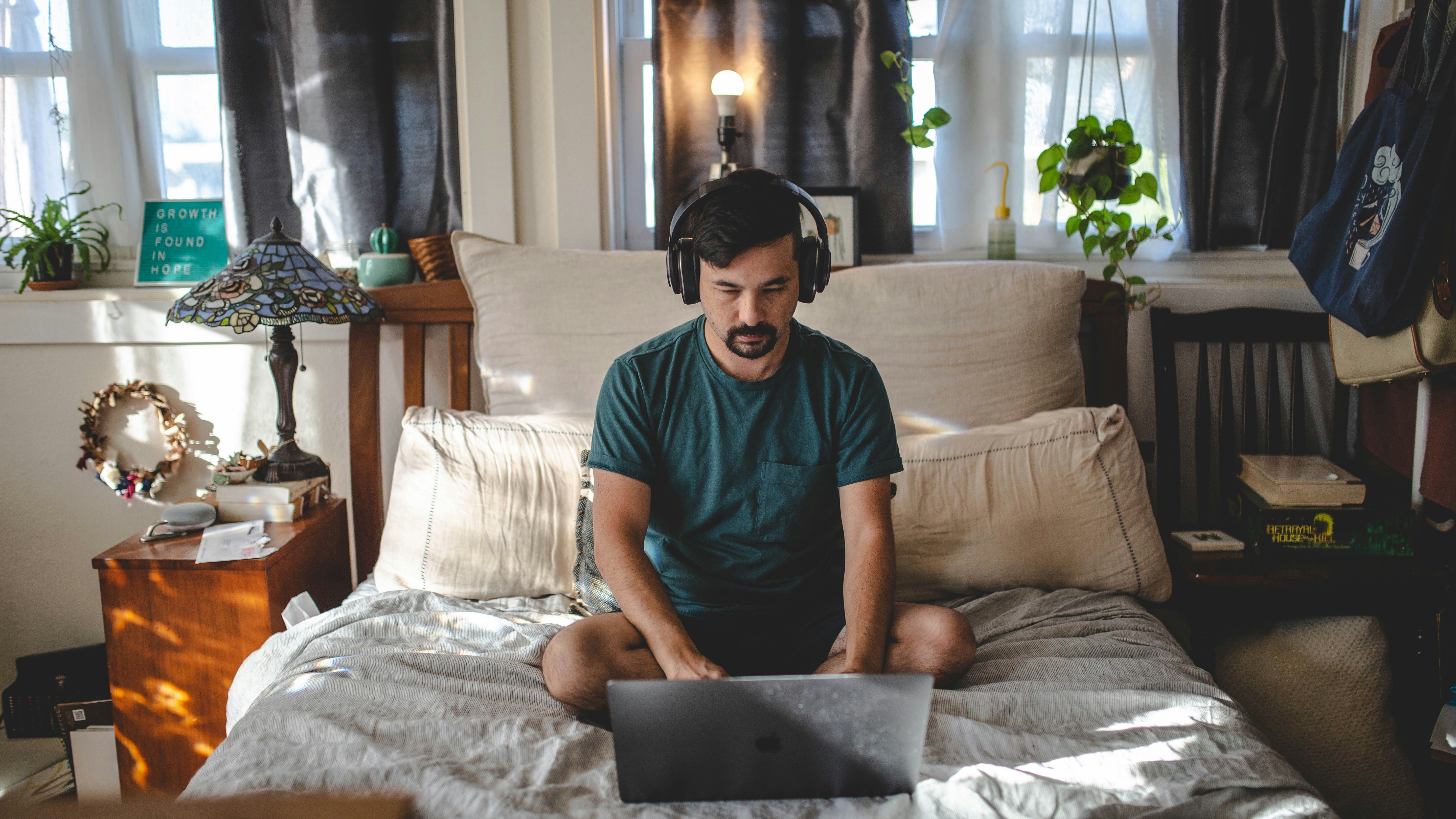 A masculine-appearing person sits cross-legged in bed looking down at a laptop, with headphones on. They are in a cluttered but cozy room, with plants, bright windows, and decorative items.