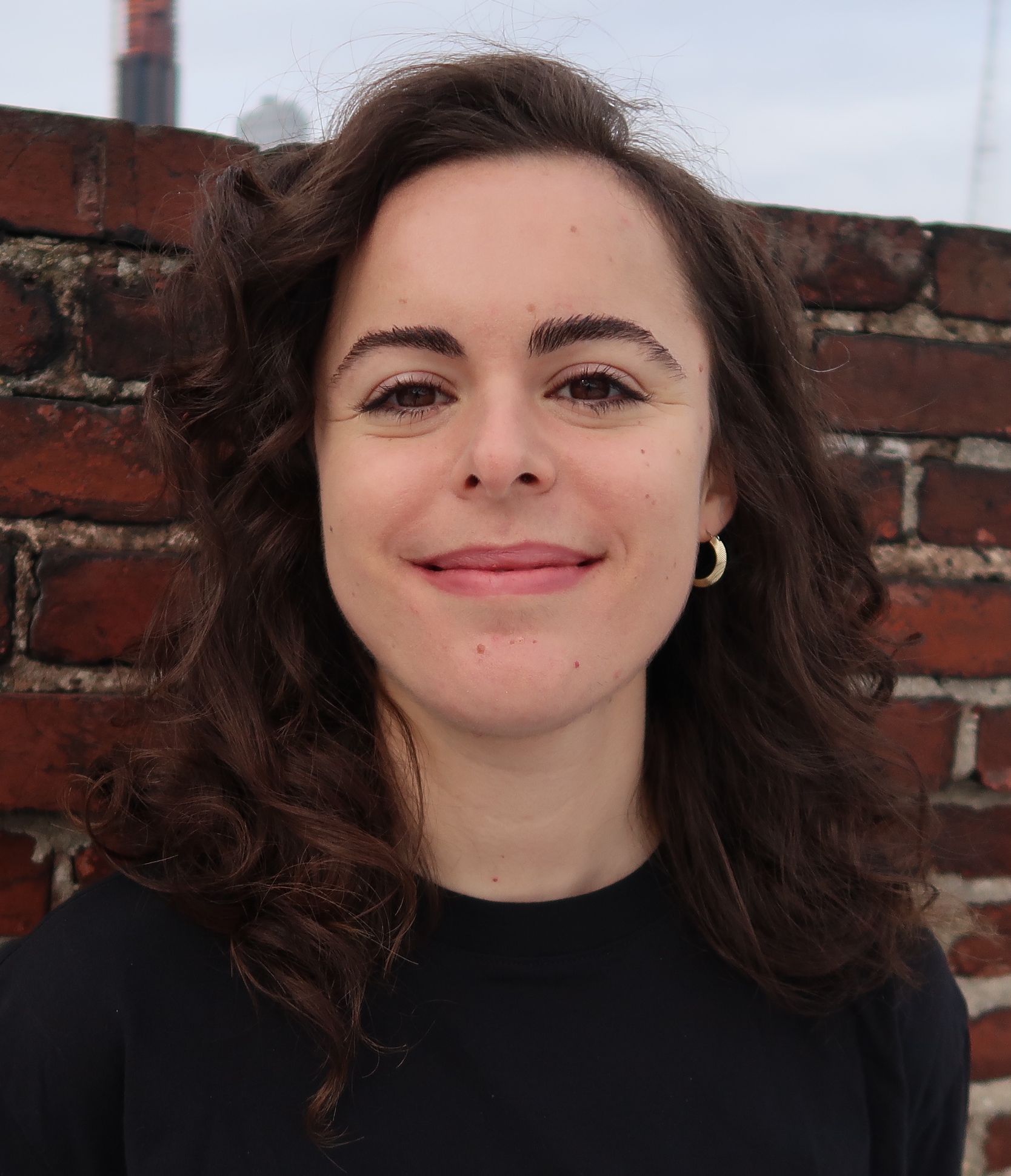 A photo of therapist and writer Elise Mendelsohn LMSW taken in Brooklyn, NY, in front of a brick wall and blue-gray sky