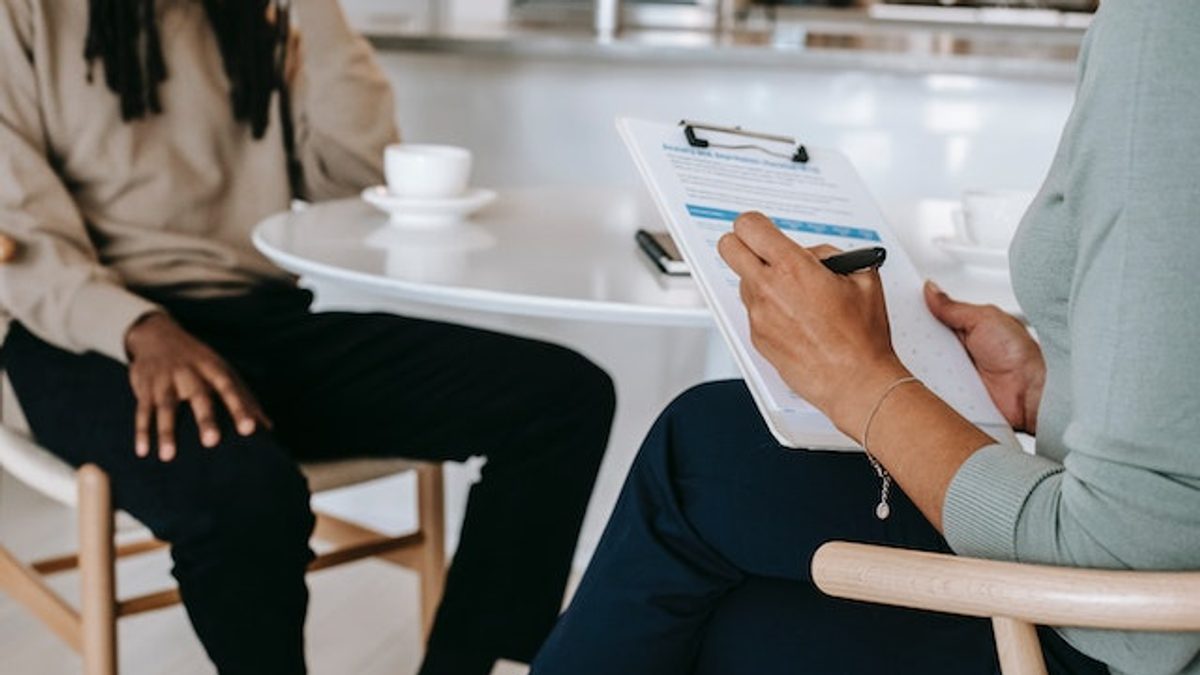 A therapist and their client seated at a café table in wishbone chairs, with the therapist taking notes on a clipboard.