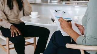 A therapist and their client seated at a café table in wishbone chairs, with the therapist taking notes on a clipboard.