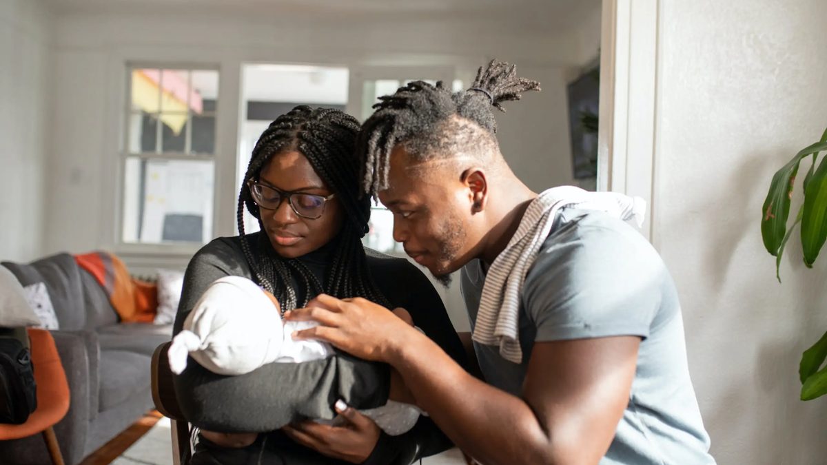 Two black parents cradling their swaddled newborn in a trendy apartment.